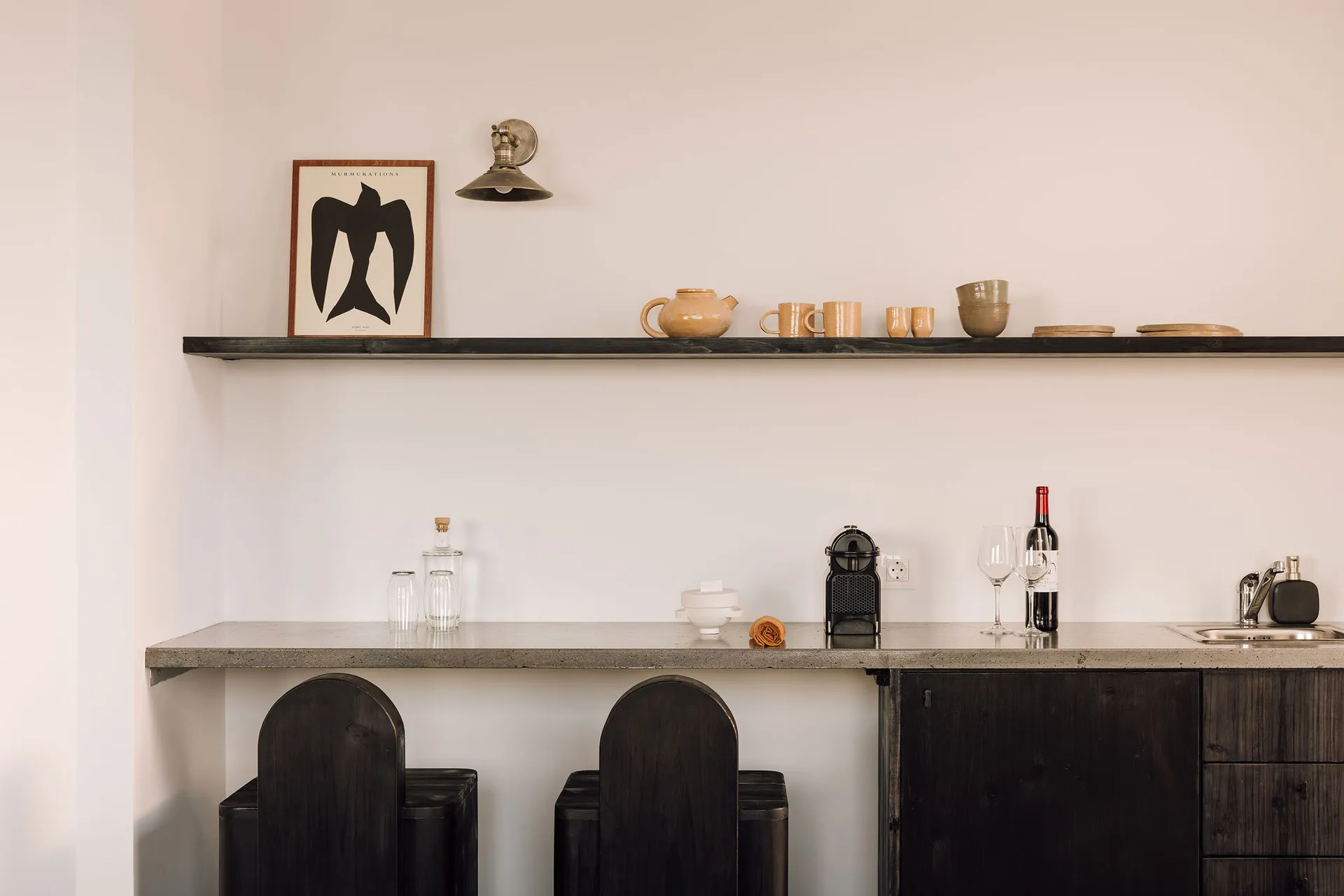 A minimalist kitchen with black stools, concrete countertop, wooden cabinets, and a wooden shelf with various ceramics and a