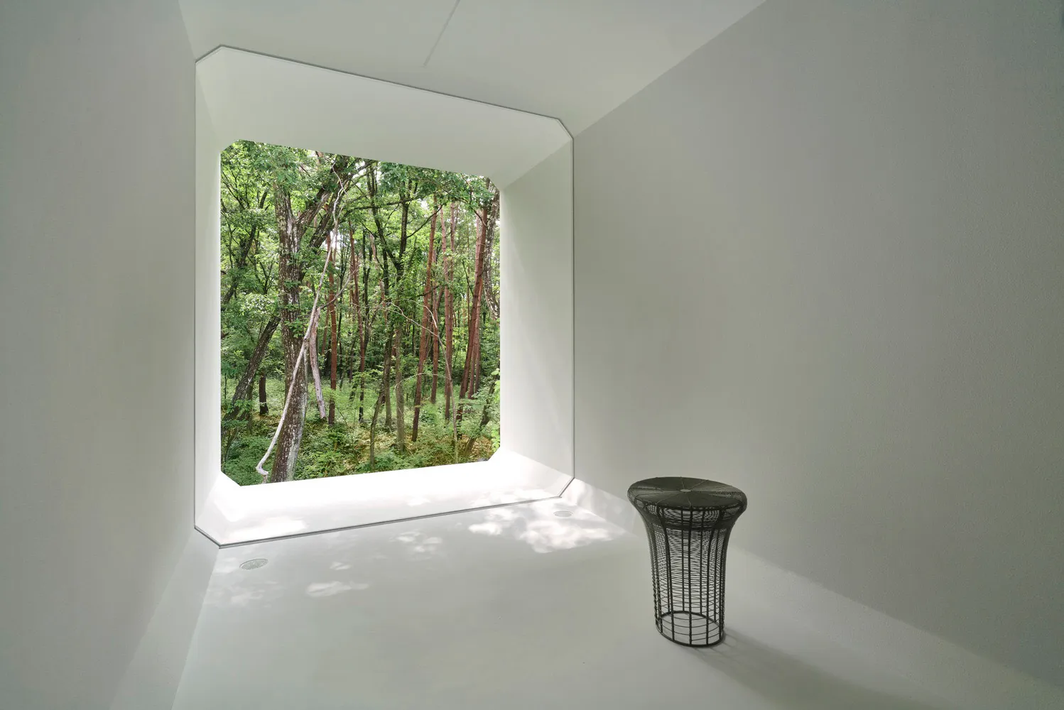 A minimalist white room with a metal stool and forest view through a large window.