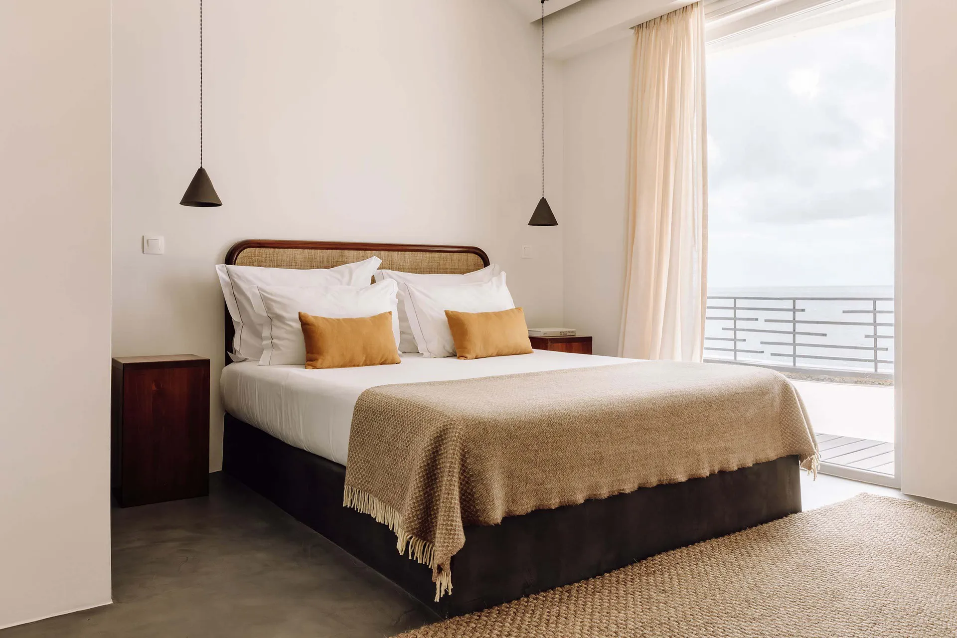 A modern bedroom with a wooden bed frame, white linens, and golden pillows, featuring a large window overlooking the sea.