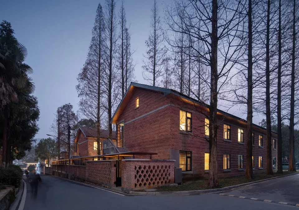 Residential building with red brick walls and illuminated windows, surrounded by tall trees in twilight.