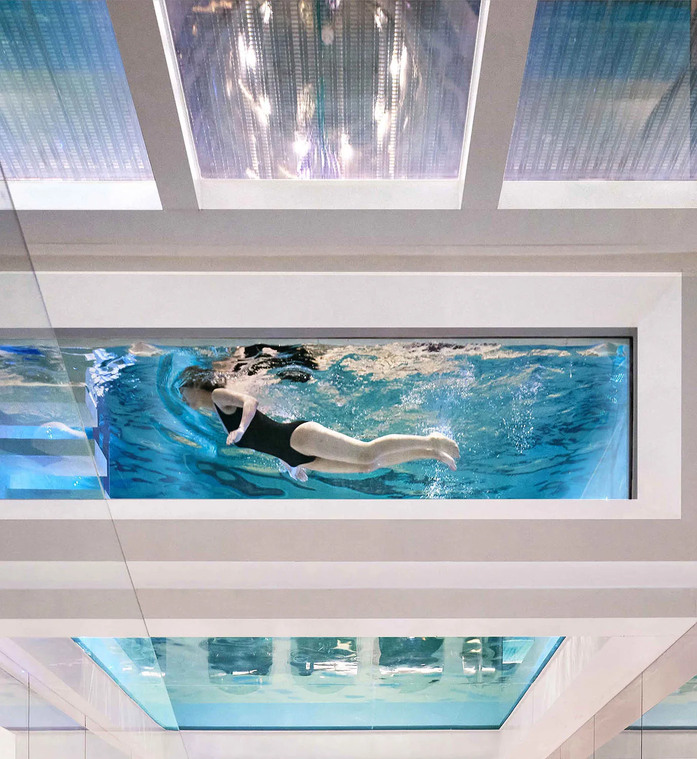 A modern glass swimming pool embedded in the ceiling, with a person swimming inside.