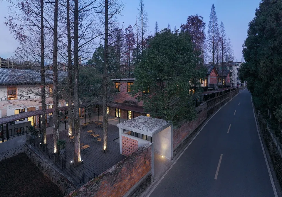 A street-side courtyard featuring brick walls, a concrete facade, and wooden decks surrounded by tall trees in a twilight set