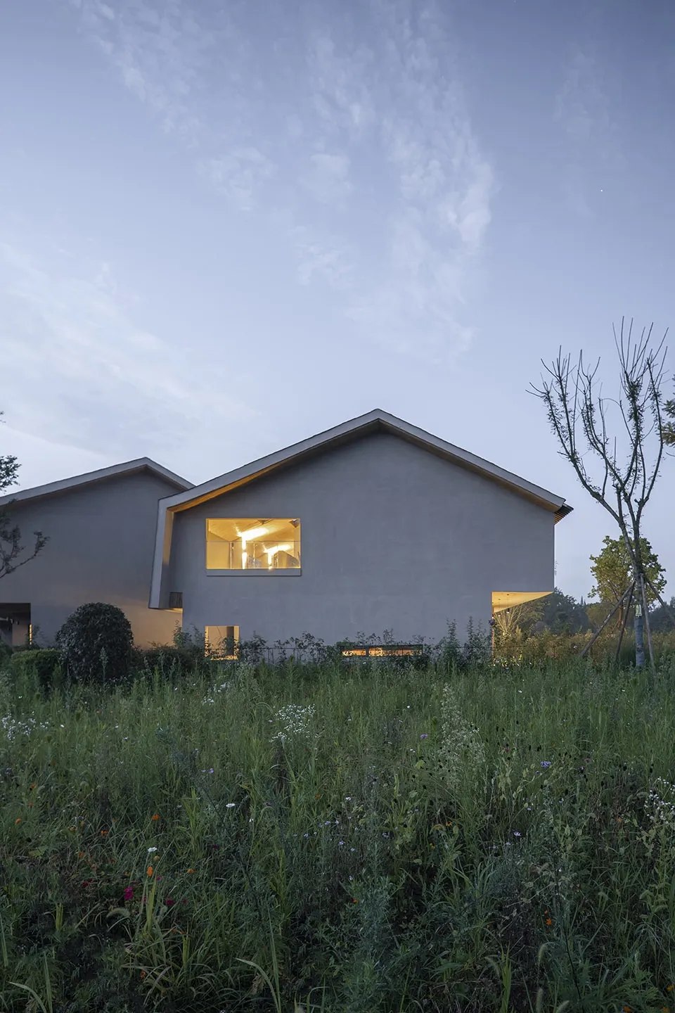 A minimalist residential concrete house with illuminated windows at dusk, surrounded by tall green grass