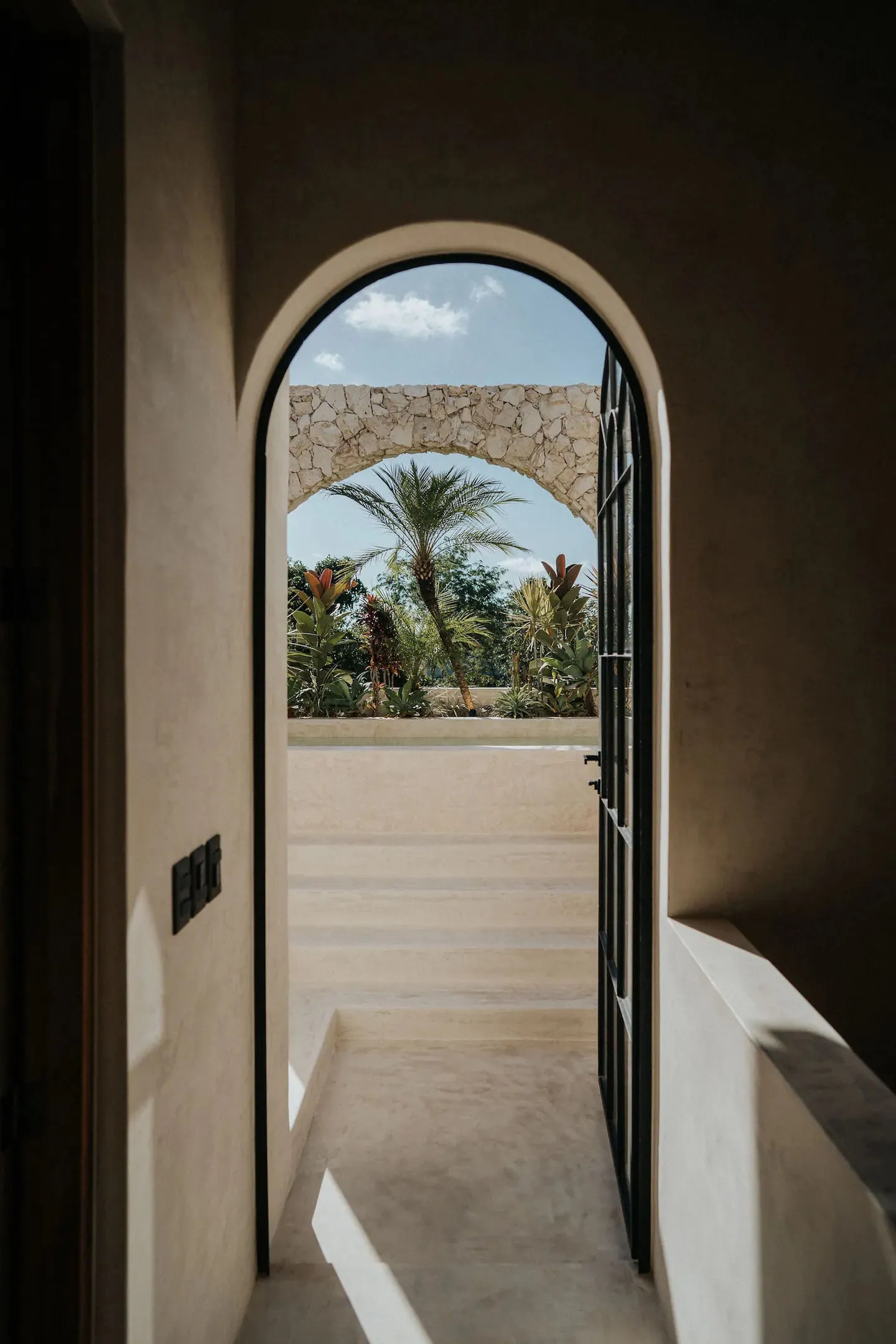 Minimalist architectural corridor with concrete floor and walls, featuring an arched doorway leading to a tropical garden.