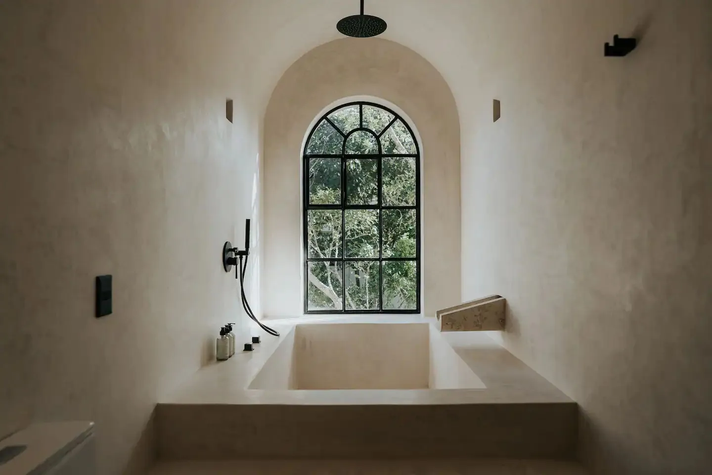 Modern bathroom featuring a concrete bathtub under a large arched window with a view of trees.