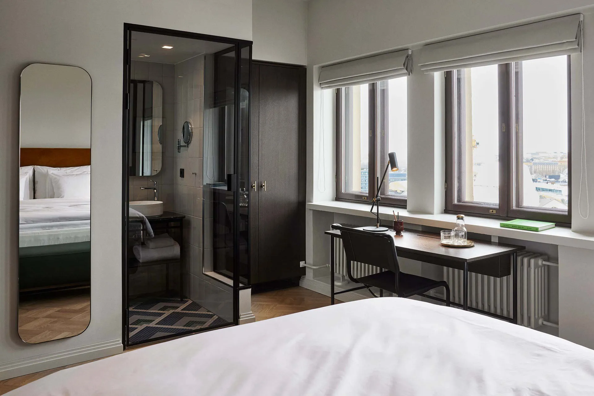 A minimalist bedroom featuring glass and wood materials, with a large bed, a desk against the window, and a bathroom visible