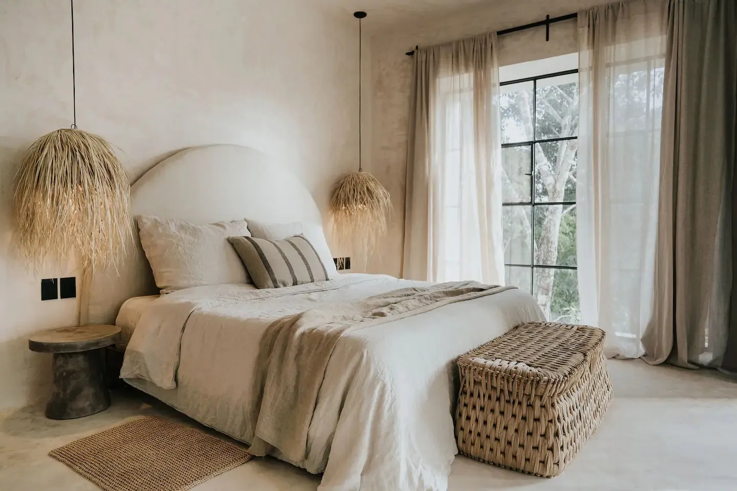 A minimalist bedroom with a textured cotton bedspread, a woven bench, and natural light from a large window overlooking green