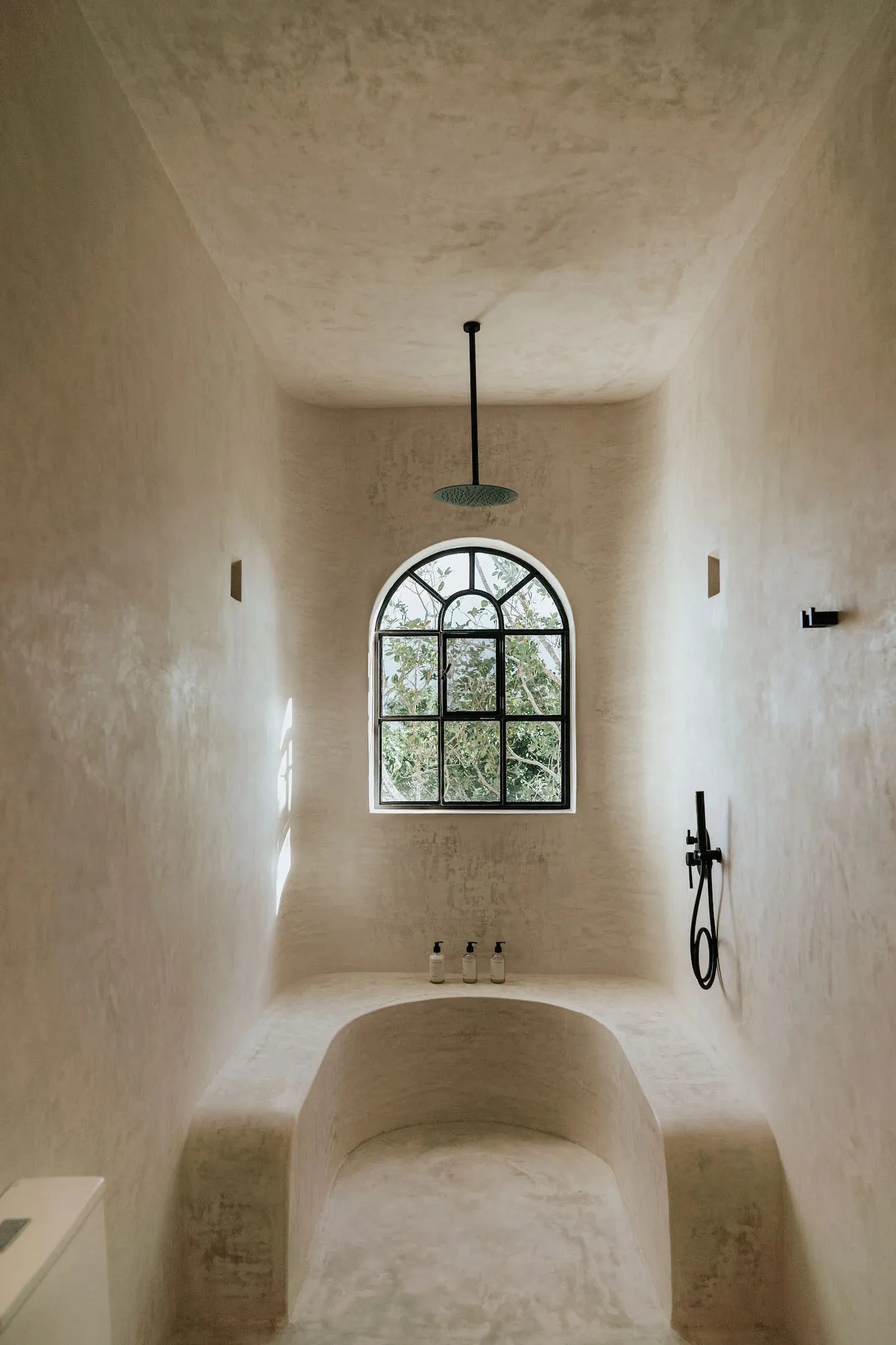 A minimalist concrete bathroom with an arched window, black fixtures, and a ceiling-mounted shower head.