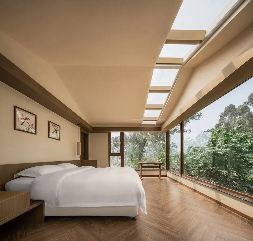 Minimalist bedroom with a white bed, wooden flooring, and large windows overlooking greenery.