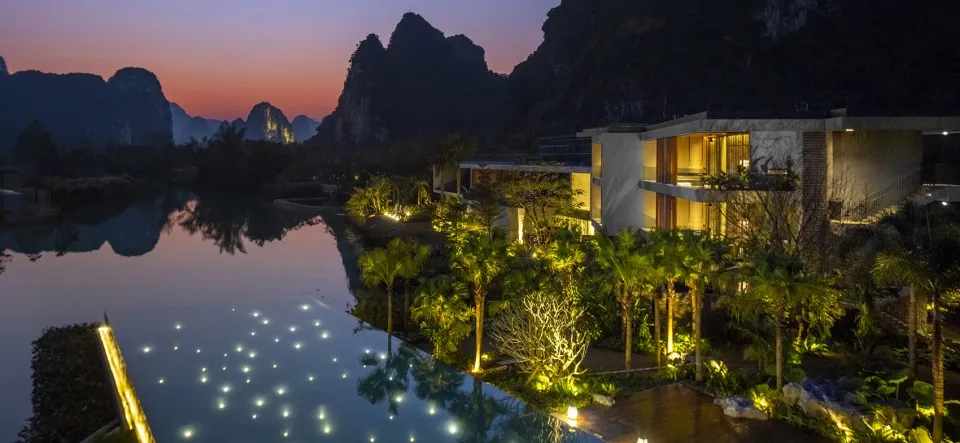 Modern resort hotel with concrete architecture, illuminated poolside area, and palm trees at dusk