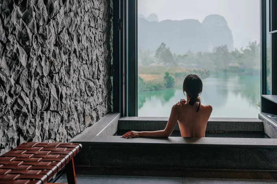 一位女士坐在石磚鋪成的SPA浴缸中,眺望寧靜湖面與遠方山景。