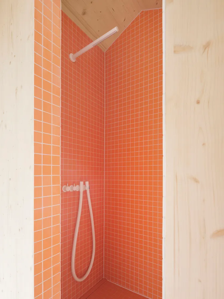A minimalist bathroom with orange ceramic tiles on the walls and floor, featuring a white showerhead and hose.