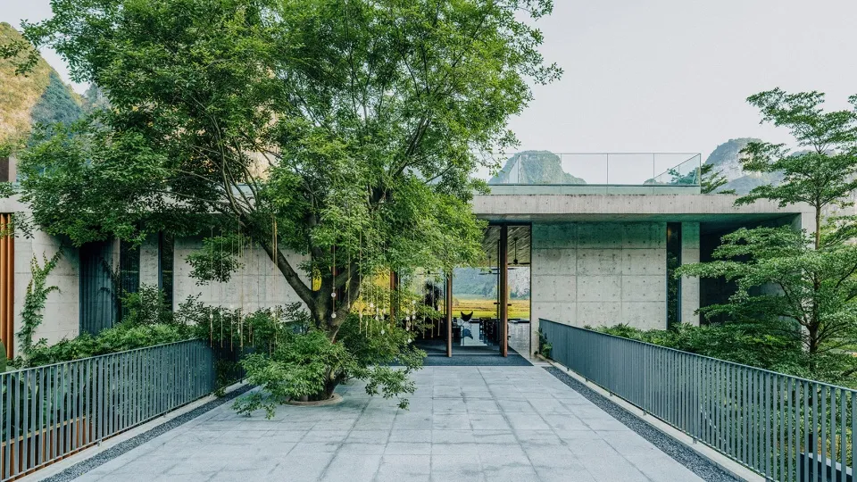 一座現代建築,擁有混凝土外牆與玻璃元素,周圍環繞綠樹與山景。