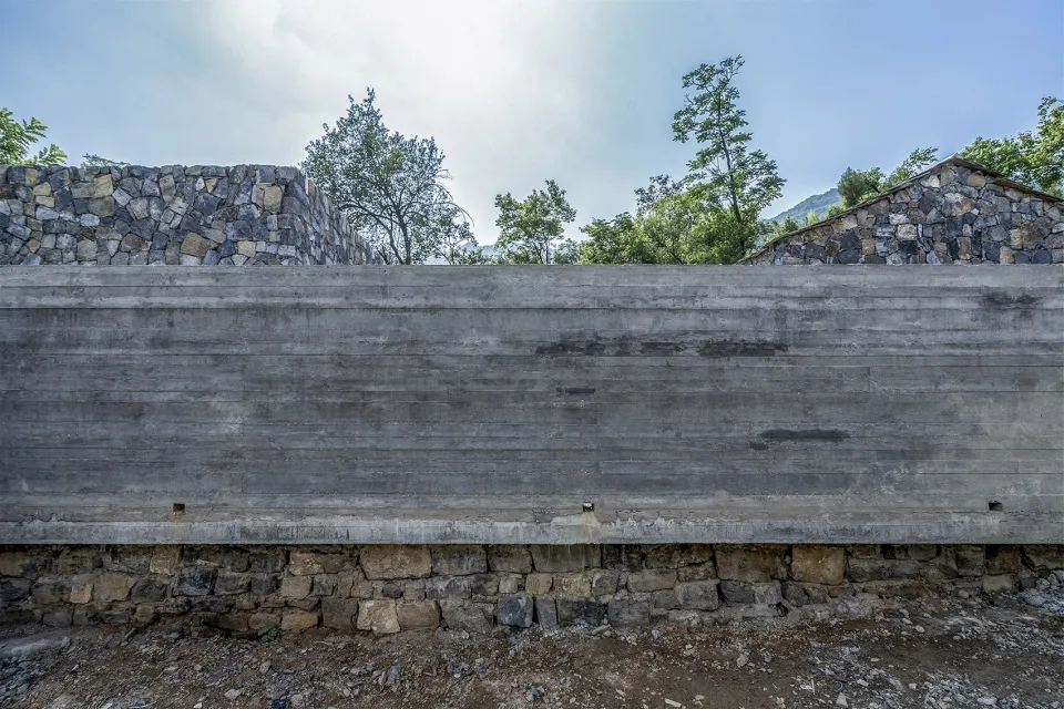 A modern concrete wall with a rough texture, set against a backdrop of green trees and a clear blue sky.