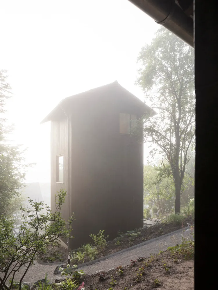 A modern minimalist house made of wood with a single window and surrounded by greenery in a foggy environment.