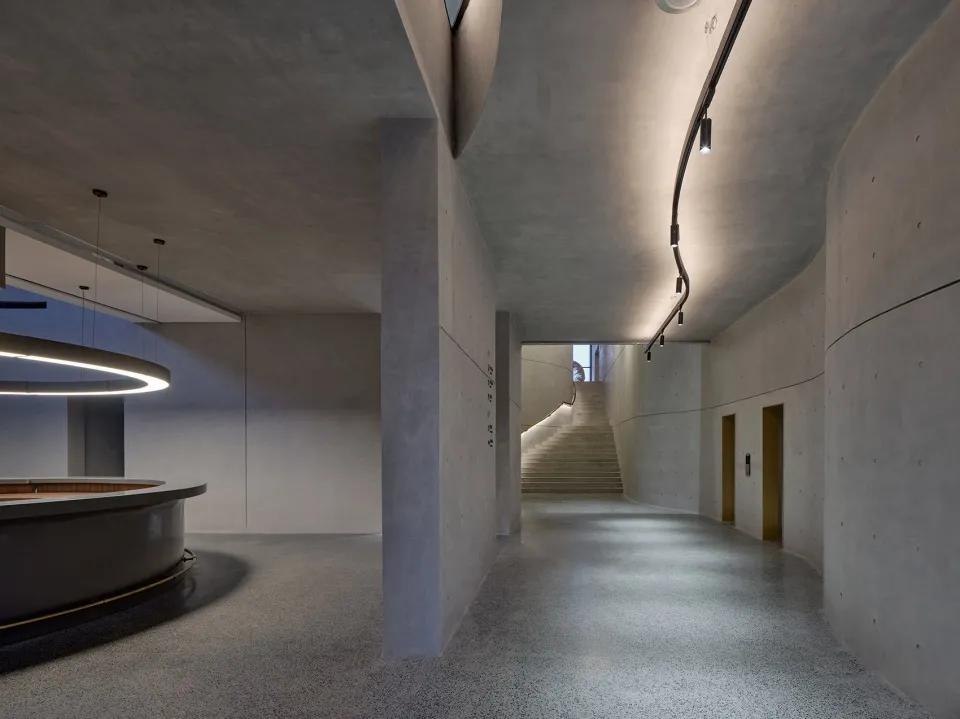 A modern office space with concrete walls and floors, featuring a curved reception desk and a staircase leading to another le