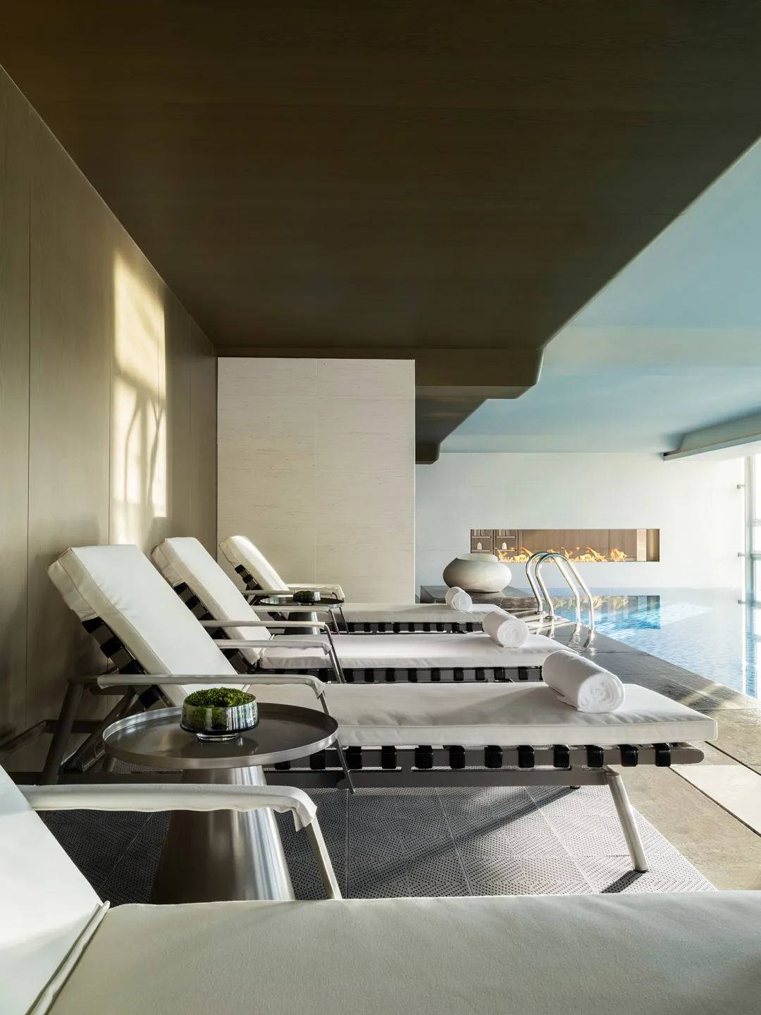 Minimalist spa lounge area with concrete walls and floor, featuring a poolside fireplace and white lounge chairs.