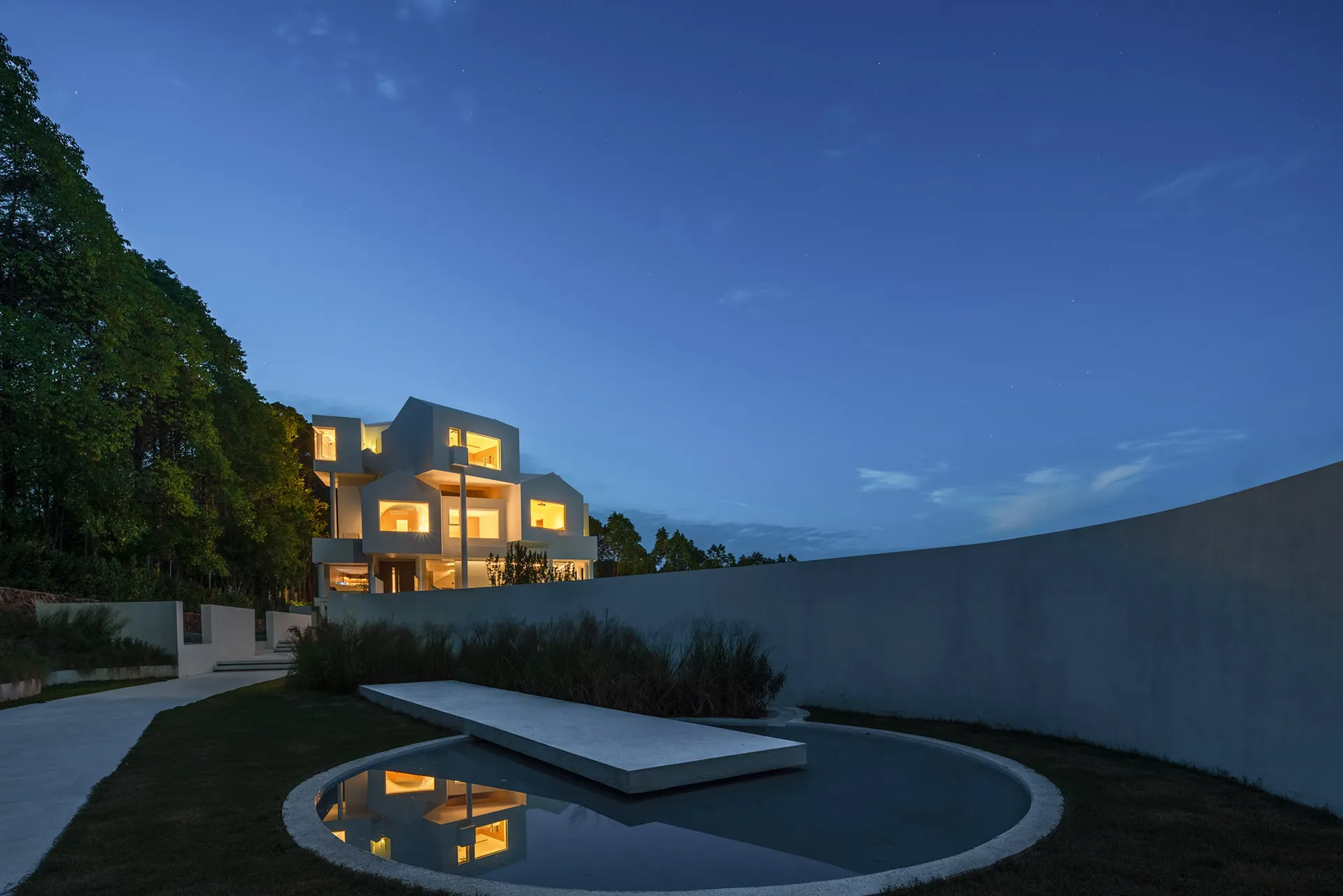 A modern, minimalist residential exterior with a courtyard and pond at dusk.