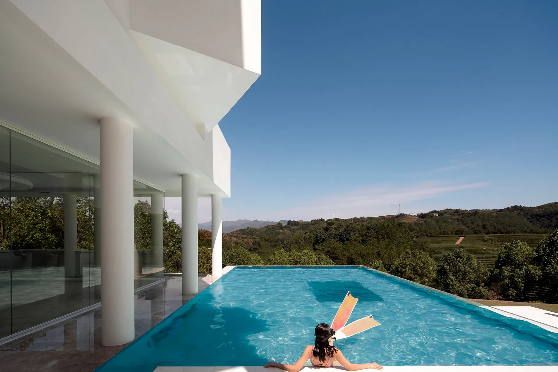 A minimalist residential home with a glass infinity pool overlooking scenic green hills under a clear blue sky.
