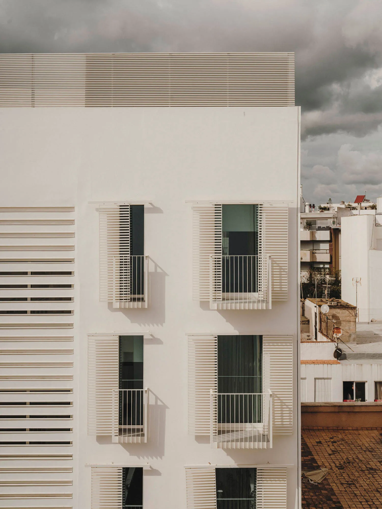 Modern residential building with white concrete exterior and white metal railings on balconies against a cloudy sky.