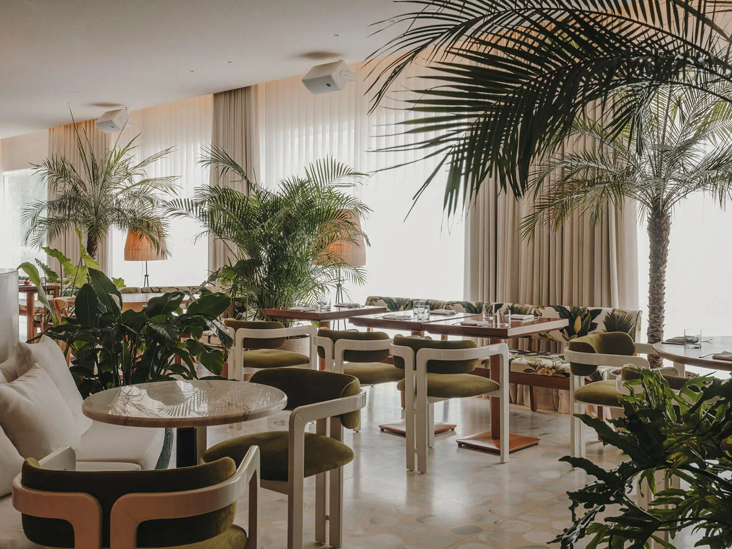A modern restaurant interior featuring greenery, wooden tables, fabric seating, glass walls, and ceramic elements.