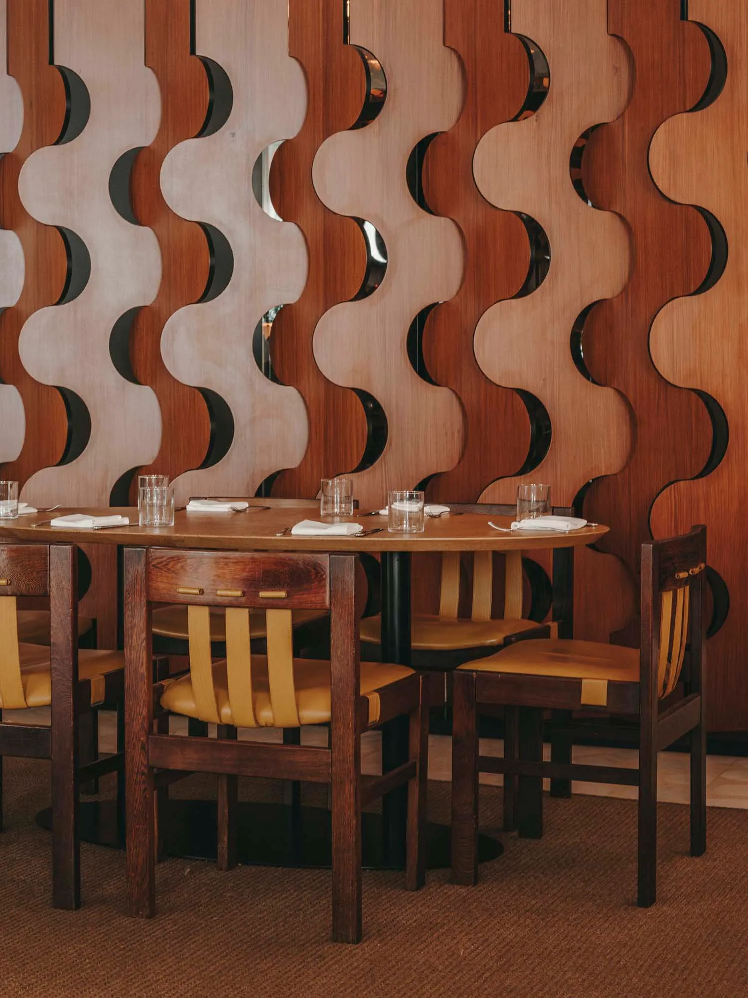 A modern restaurant interior with a wooden design featuring a round table, yellow chairs, and a decorative wave-patterned wal