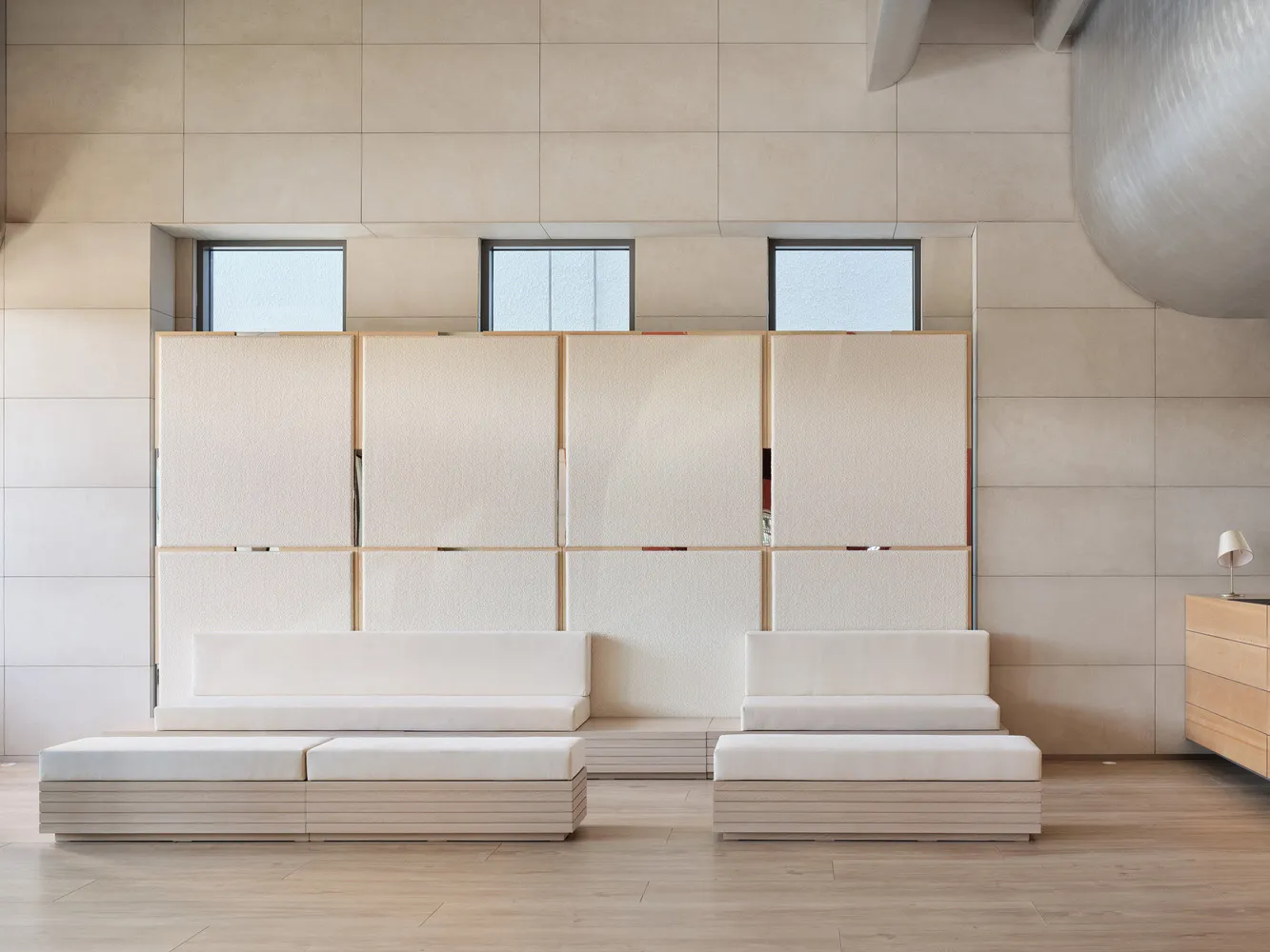 Minimalist living room with white sofas and wooden bench seating, glass windows, and a modern light fixture.