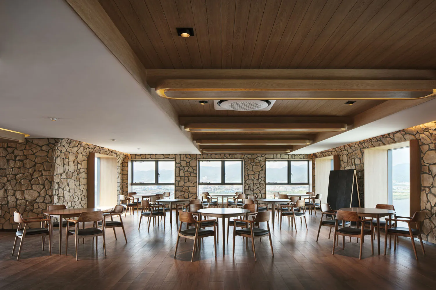 A spacious, modern restaurant cafe with round tables, wooden chairs, and stone walls, featuring a warm and inviting atmospher