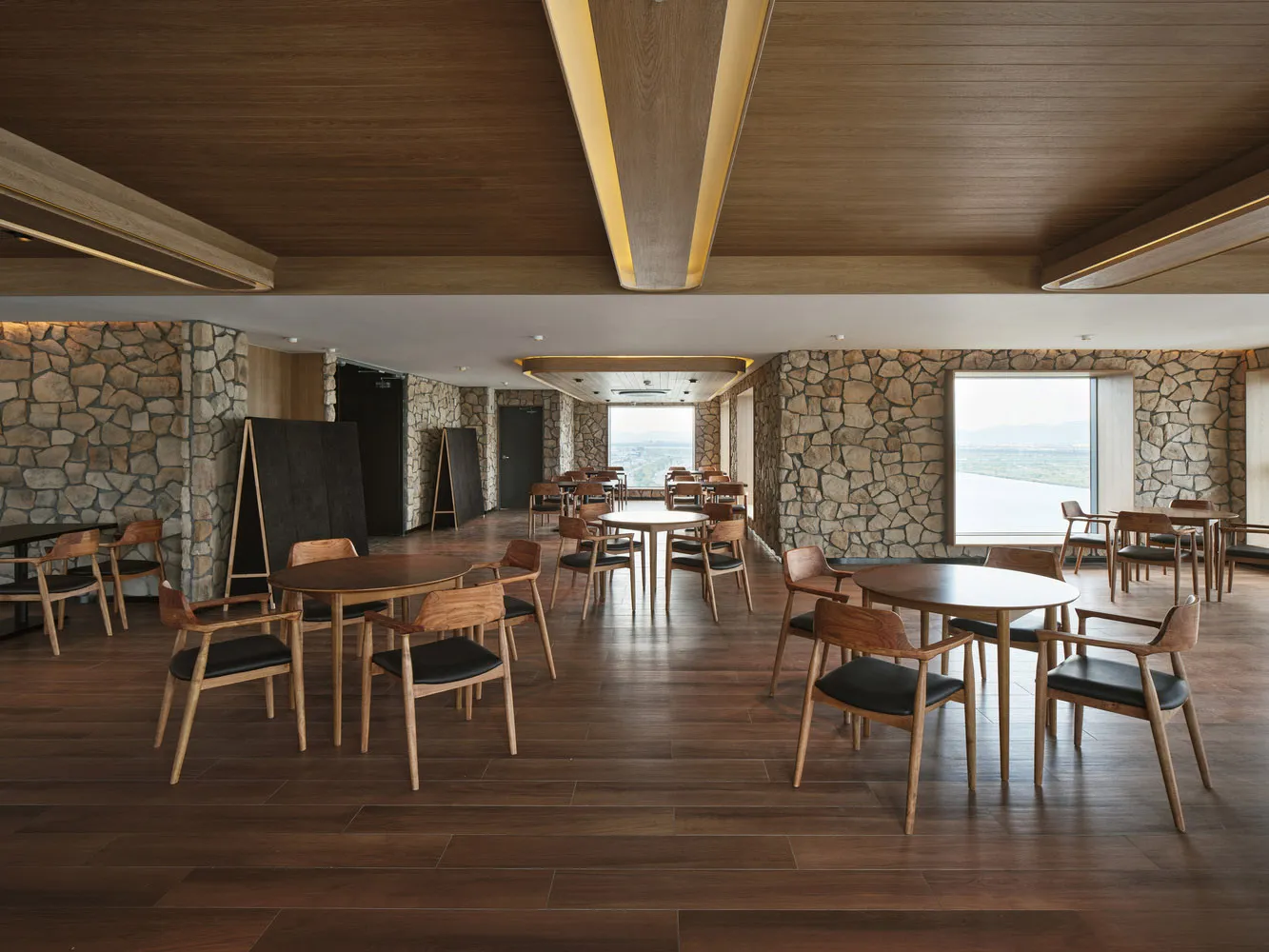 Interior of a restaurant cafe with wooden tables and chairs, stone accents, and large windows.