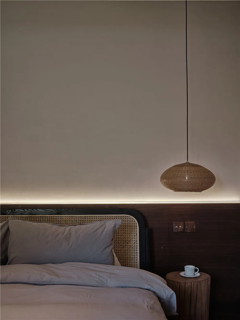 A minimalist bedroom with a woven wicker pendant light and a bed adorned with grey pillows and blanket, set against a plain w