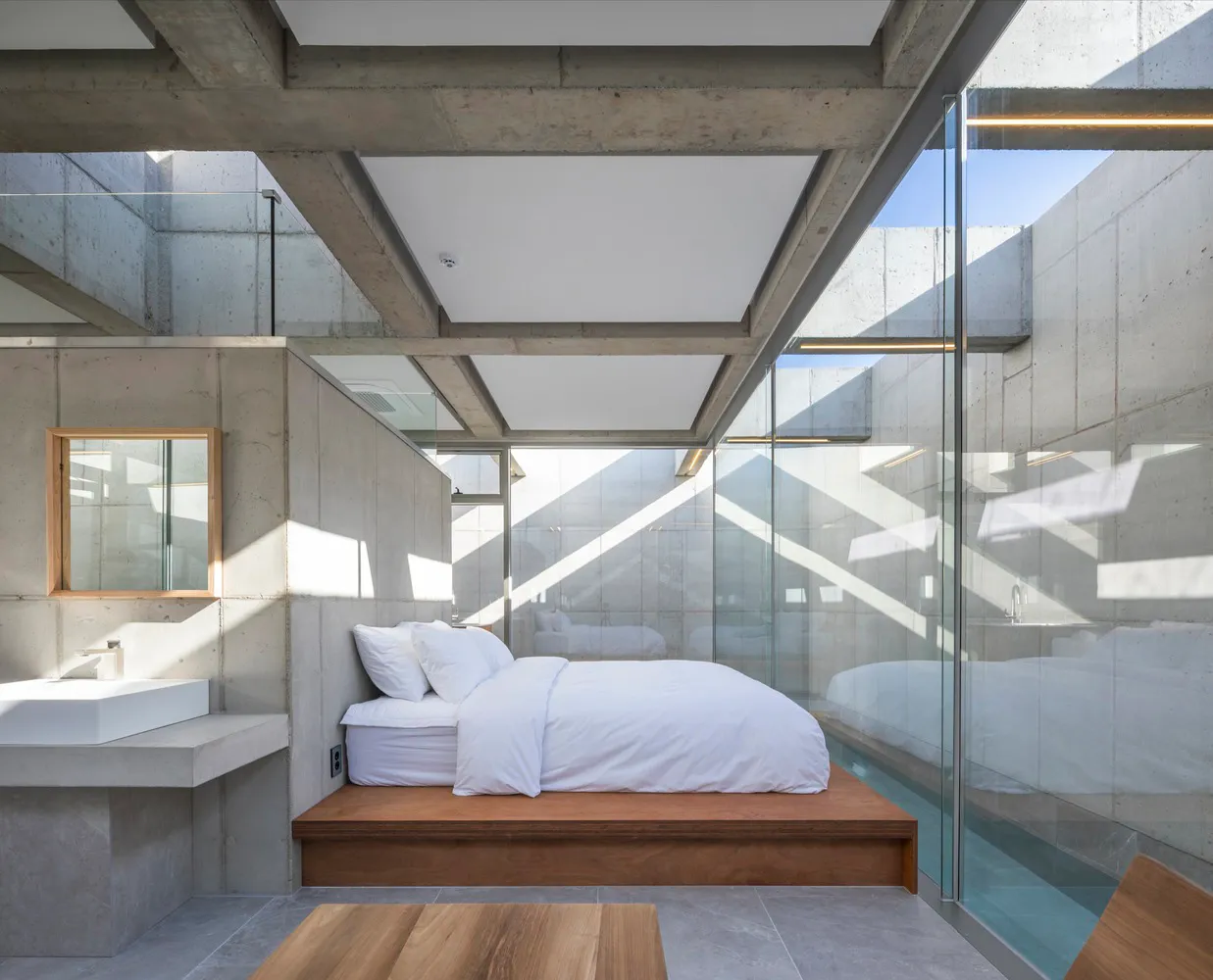 Modern minimalist bedroom with concrete walls, a raised bed platform, and a glass wall showcasing a pool.