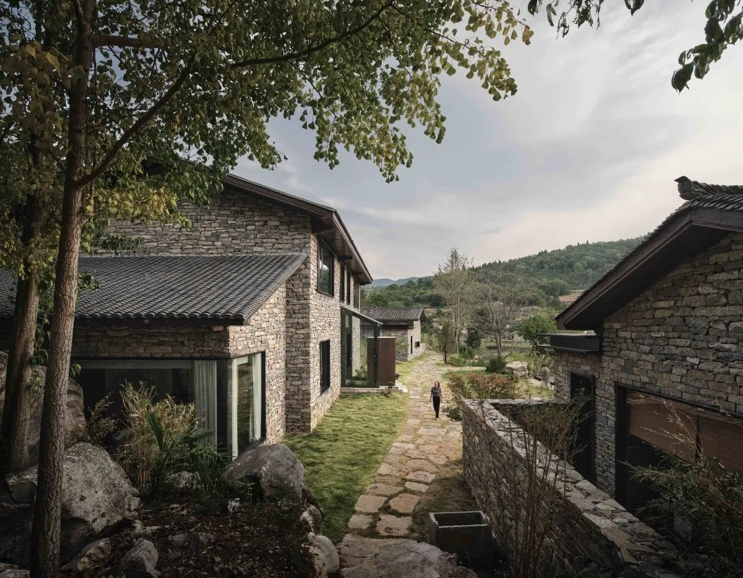 A stone and glass exterior of a country house with a gravel path, green grass, and trees.