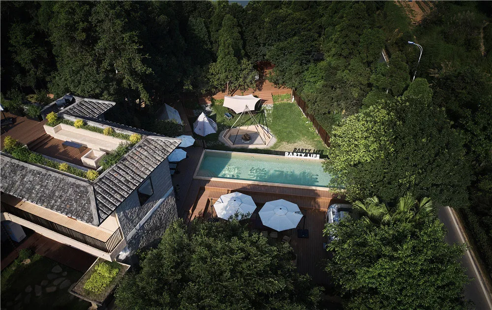 Aerial view of a modern residential outdoor space with a pool, umbrellas, and wooden decks surrounded by lush greenery.