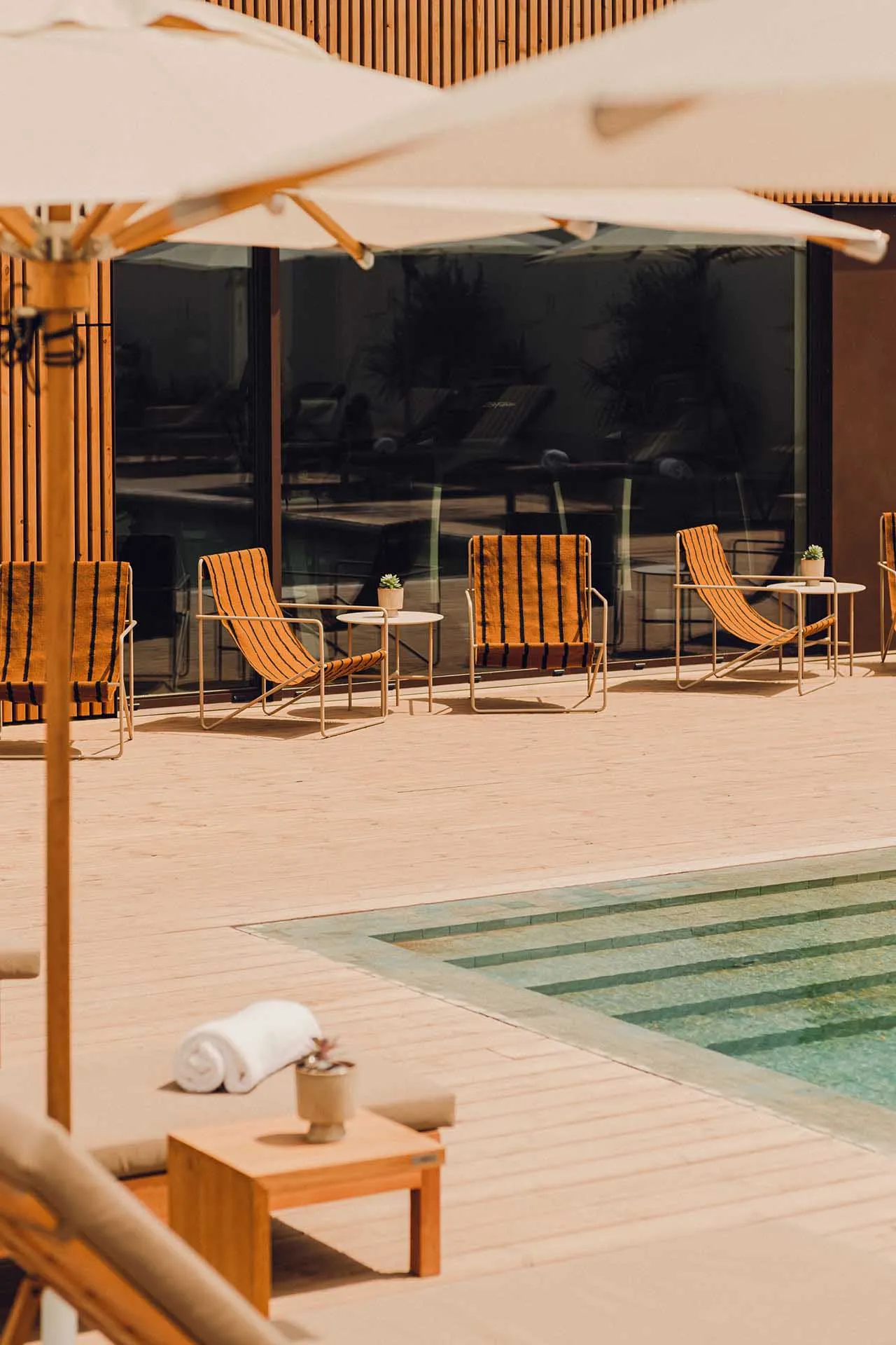 A minimalist poolside lounge area with wooden lounge chairs, glass tables, and umbrella shades.