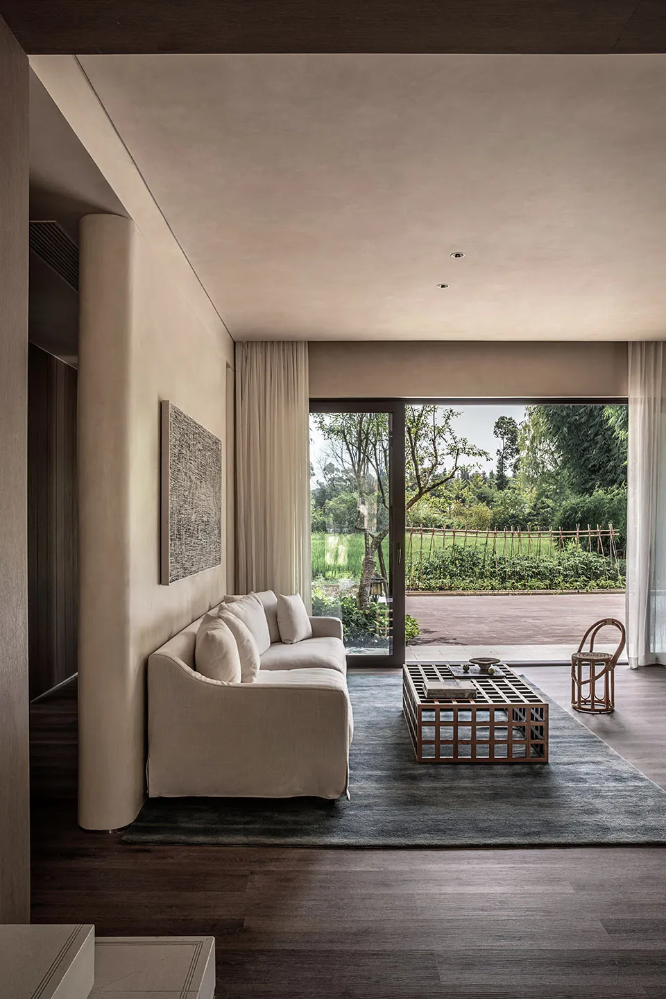 A modern living room with a beige sofa, a wooden coffee table, and a large window overlooking a green outdoor area.
