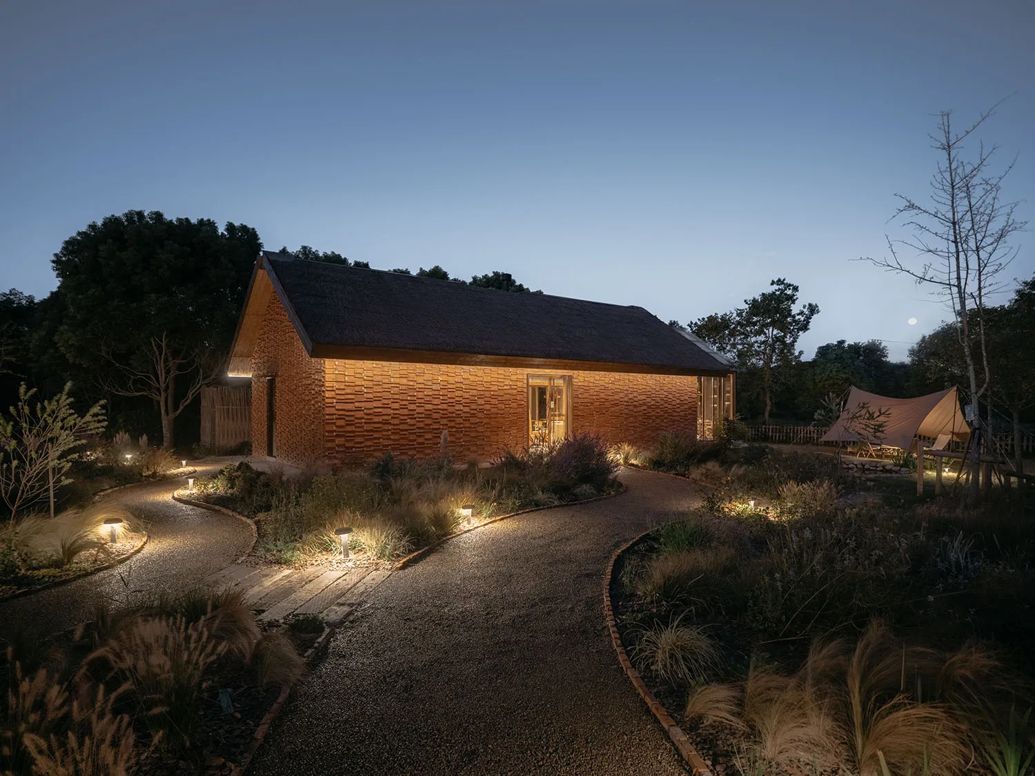 A wooden house with bamboo elements illuminated at night, featuring a curved pathway and surrounding garden.