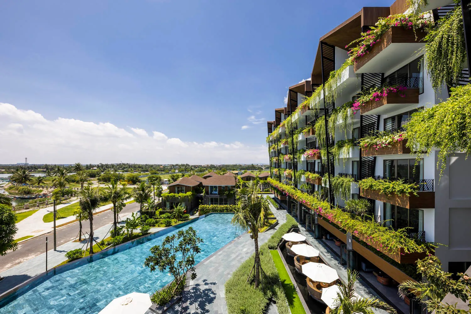 A modern hotel with concrete and glass exteriors, featuring a large pool area and lush greenery.
