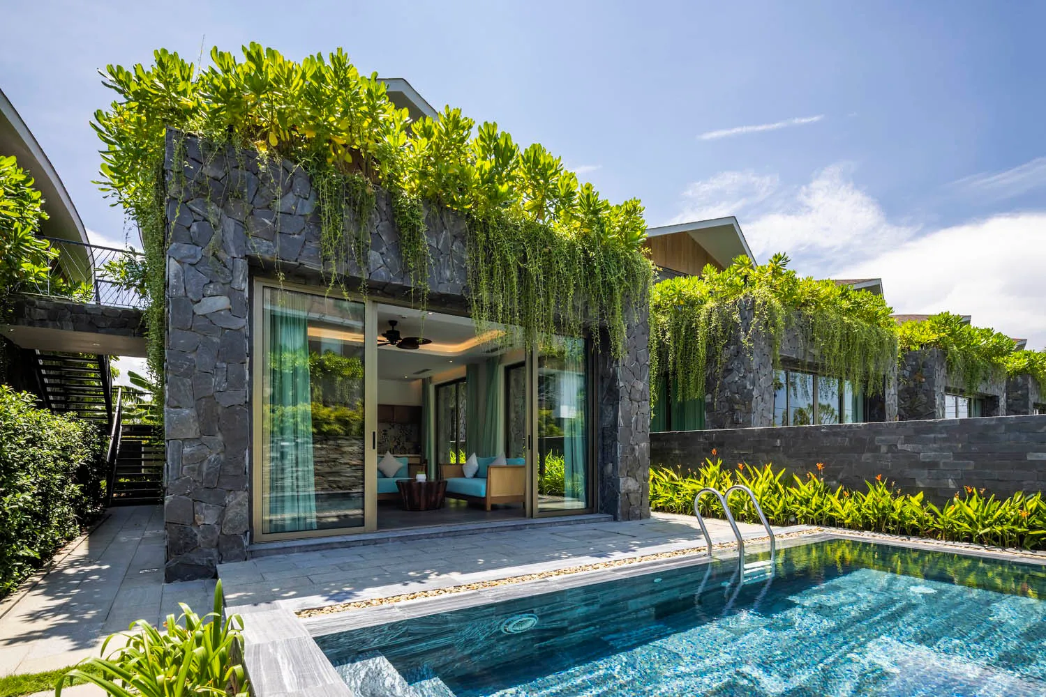 A modern villa exterior with stone walls and green leafy plants surrounding a private pool.