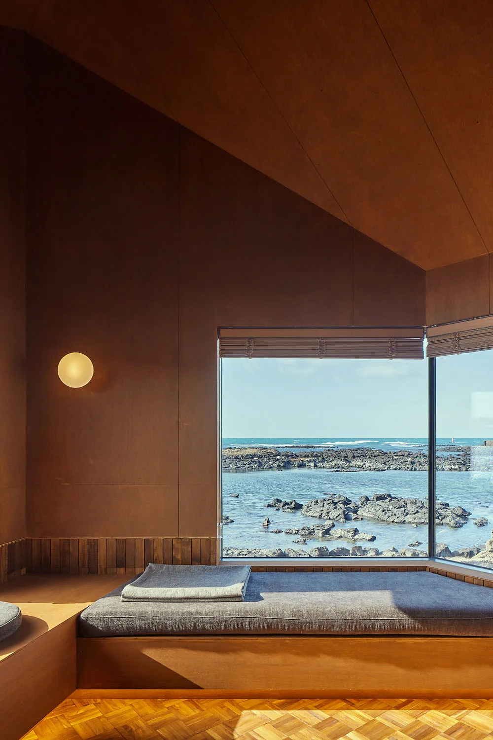A minimalistic interior with a large window overlooking the ocean, featuring wooden flooring and upholstery.