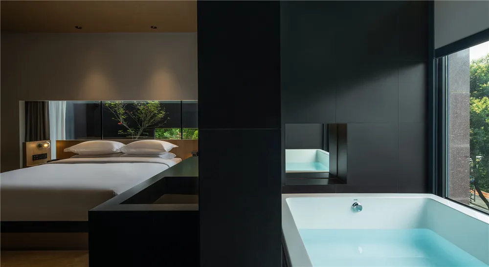 Modern, minimalist hotel bedroom featuring a sleek, black-walled bathroom with a soaking bathtub and a bed adorned with plush