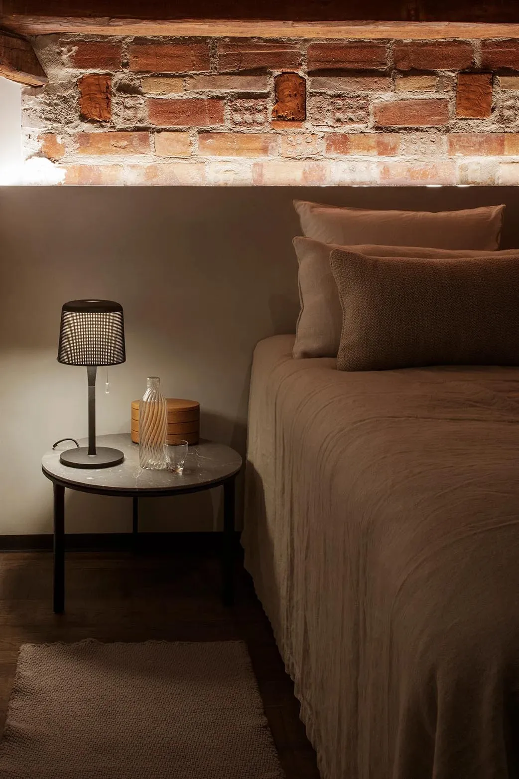 A minimalist bedroom with a bed dressed in fabric bedding and a side table with a lamp and a glass.
