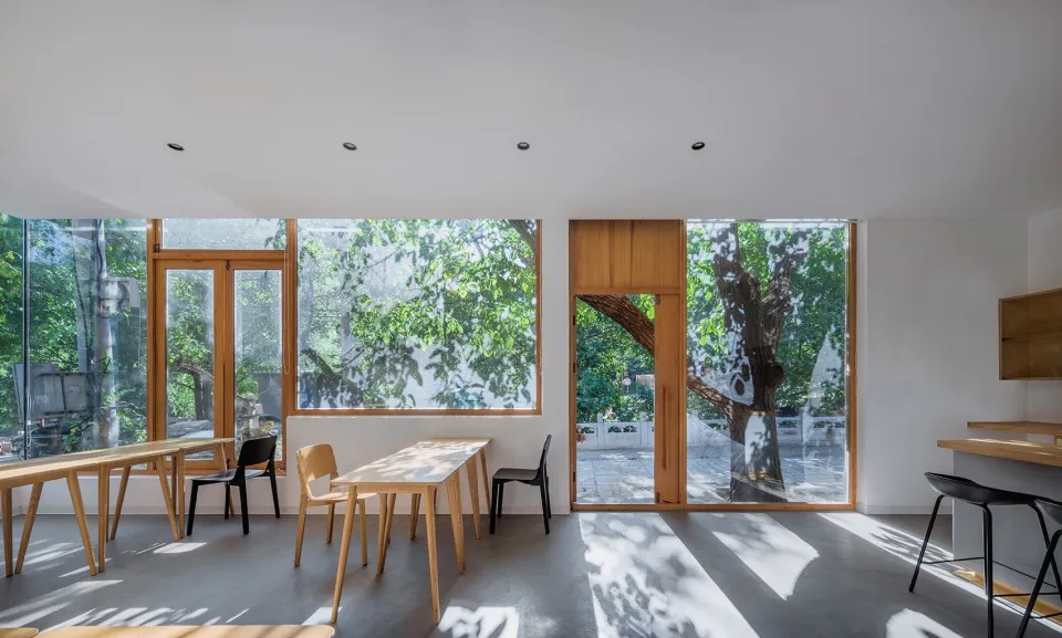 A minimalist cafe interior featuring glass walls, wooden tables, and concrete flooring.