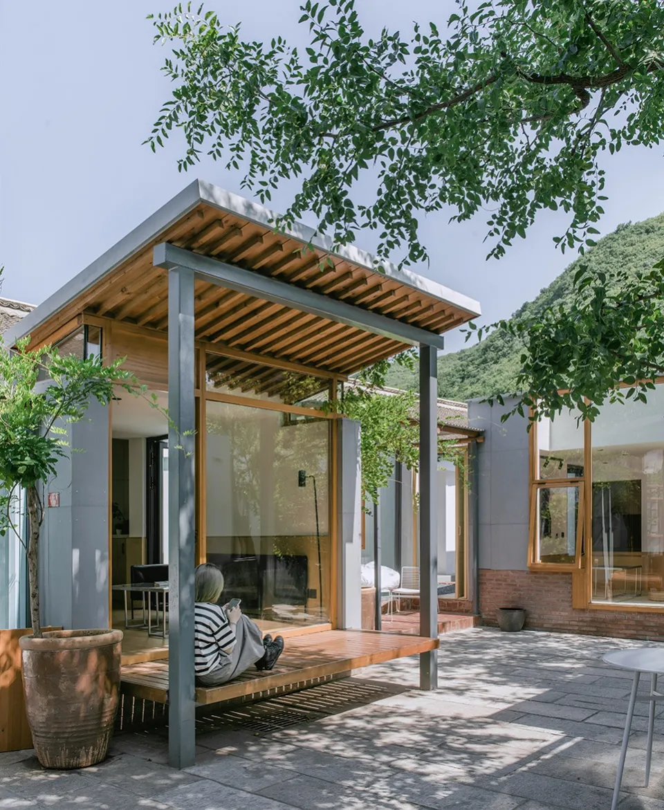 A modern minimalist exterior featuring a wooden structure with large windows, surrounded by greenery and mountains.