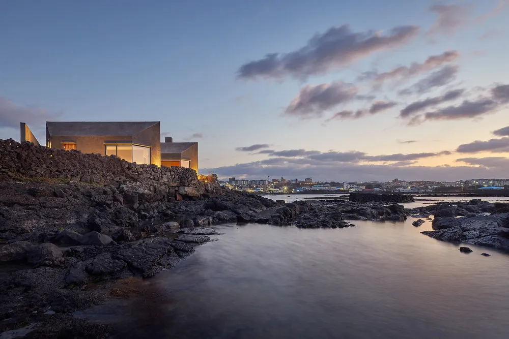 Modern house with large windows and concrete walls overlooking the water at dusk.