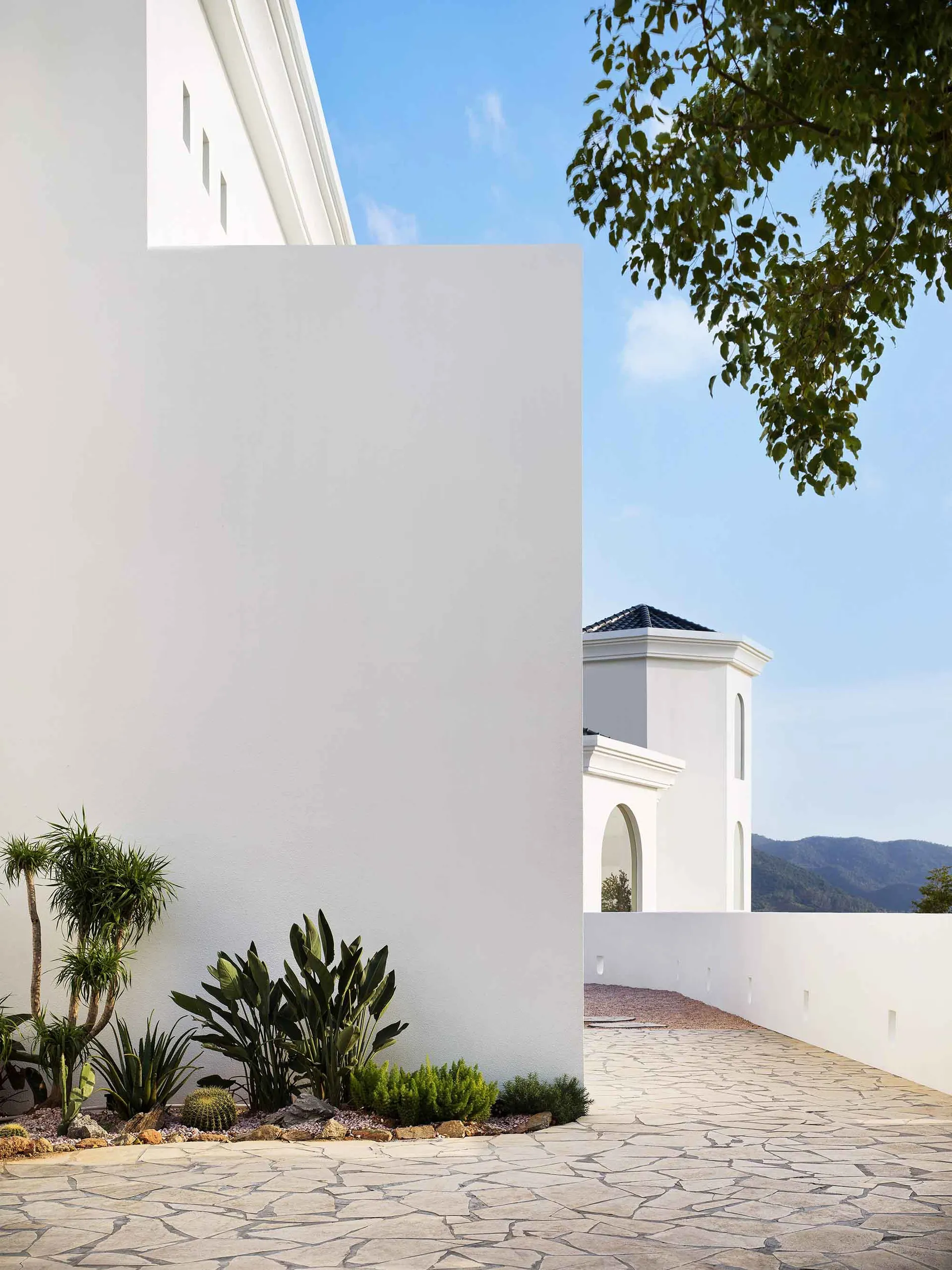 Modern exterior architectural space featuring concrete walls and a tiled pathway with greenery and a mountainous backdrop.