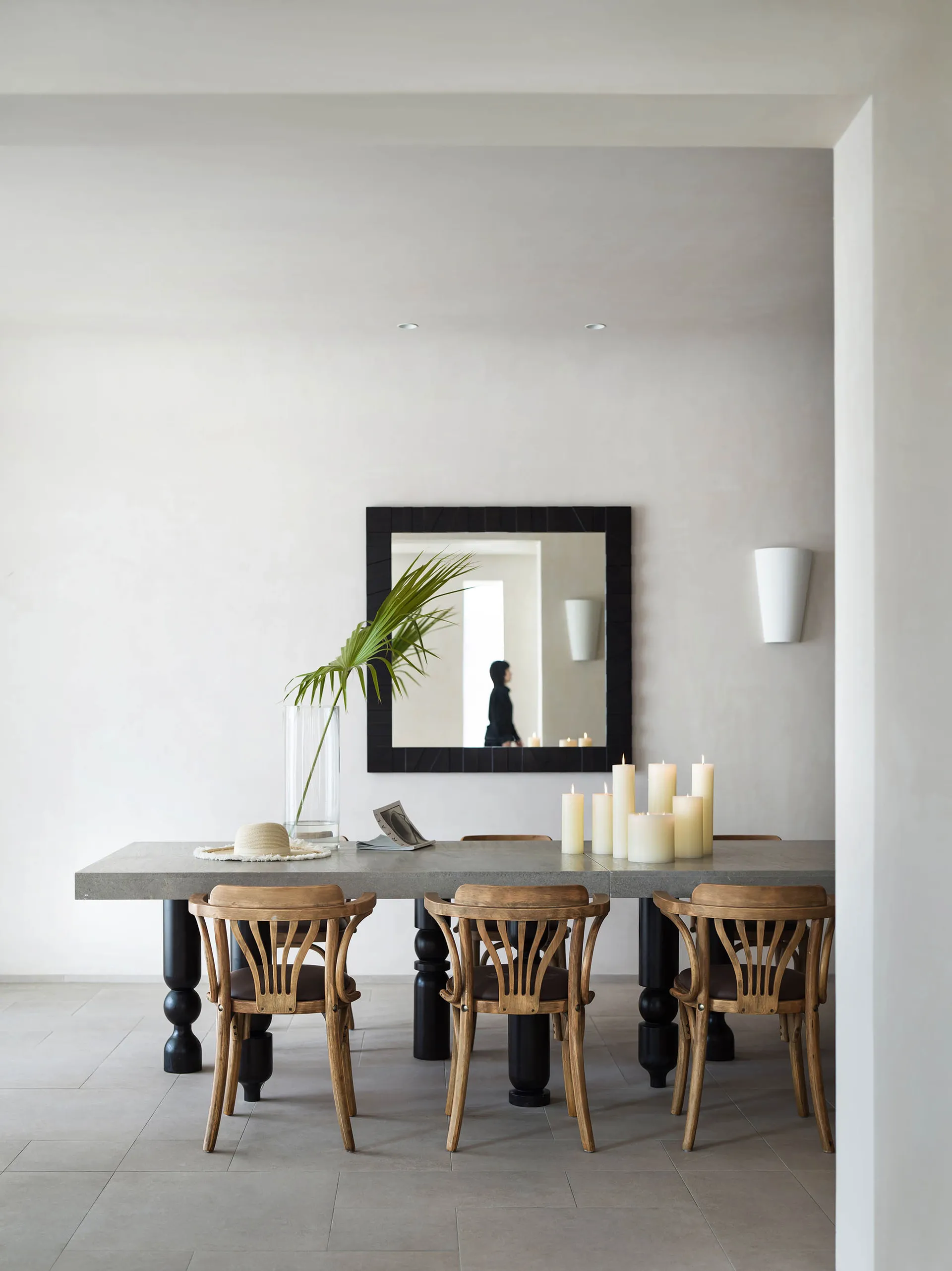 A modern minimalist dining room featuring a concrete table and wooden chairs, with a large mirror on the wall, candles, and a