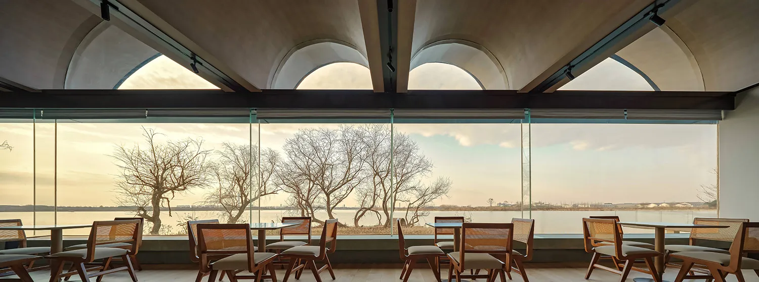 Interior view of a modern restaurant featuring large glass windows, concrete columns, and wooden tables and chairs.