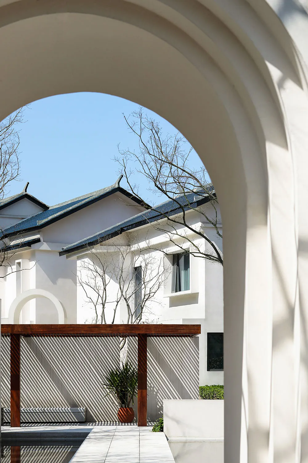 A white plaster residential space with a wooden pergola and potted plants under an archway.