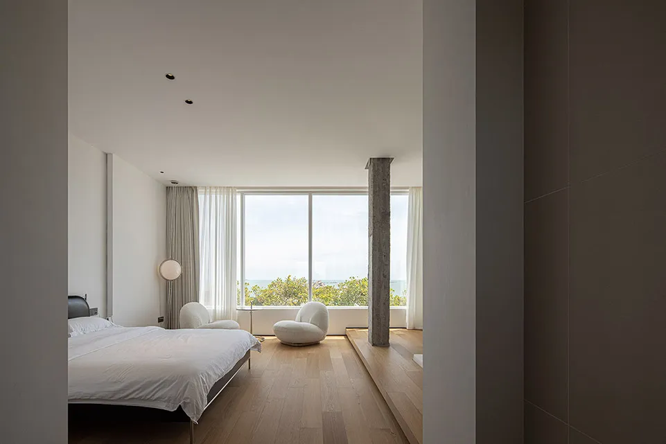 A modern bedroom with a white bed, white chairs, and wooden flooring, featuring a large window with curtains and a concrete c