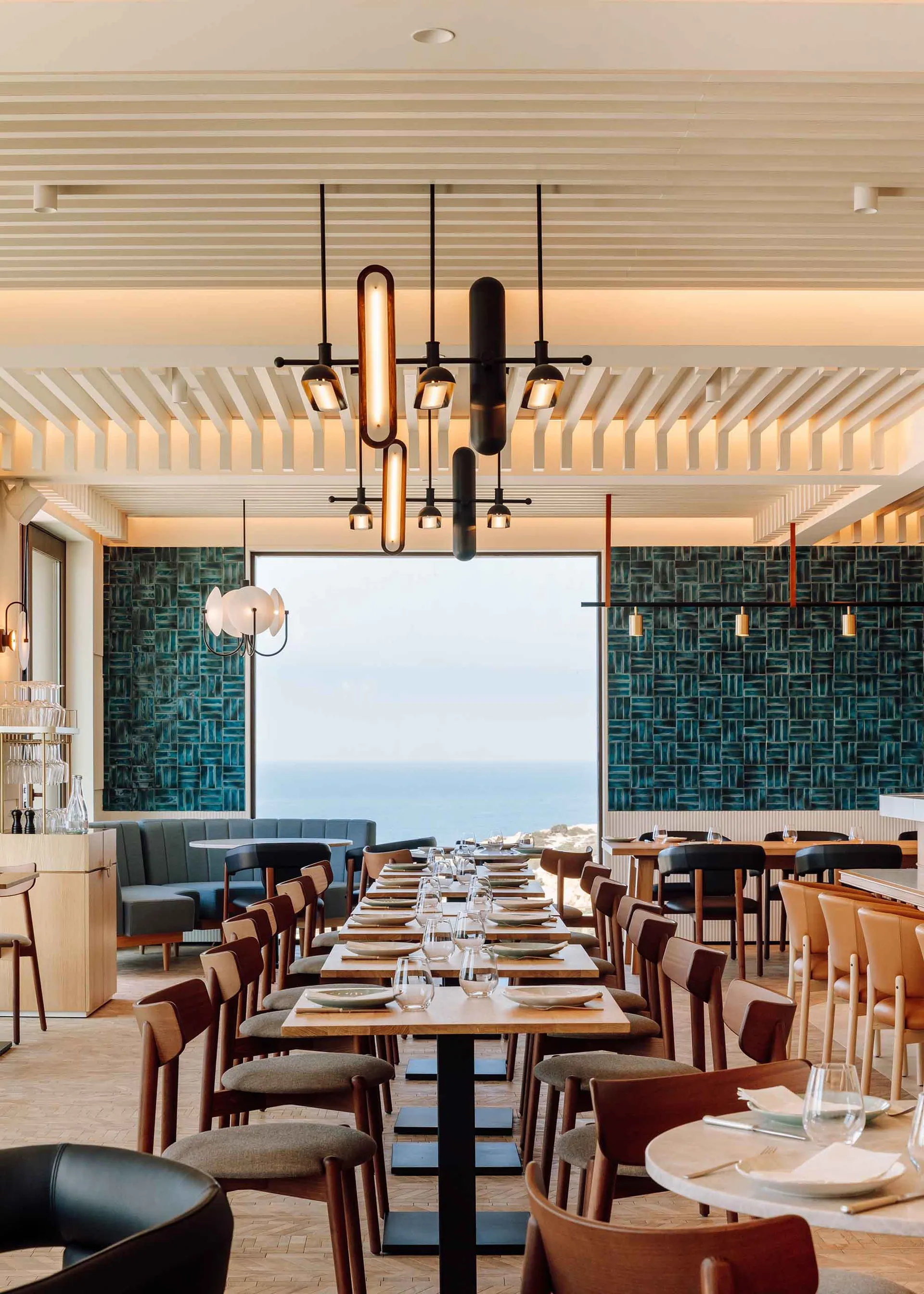 Modern restaurant interior featuring wooden tables, glass chandeliers, and blue-tiled walls with a view of the ocean.